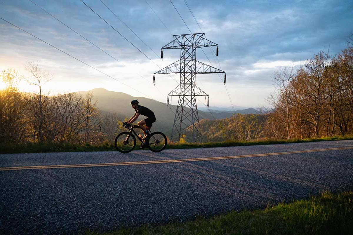 Cyclist riding down road with electrical line, trees and mountains behind.